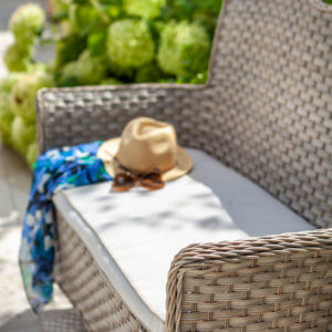 close up of Hartman heritage 2 seat rattan garden bench- beech/dove with hat and sunglasses on