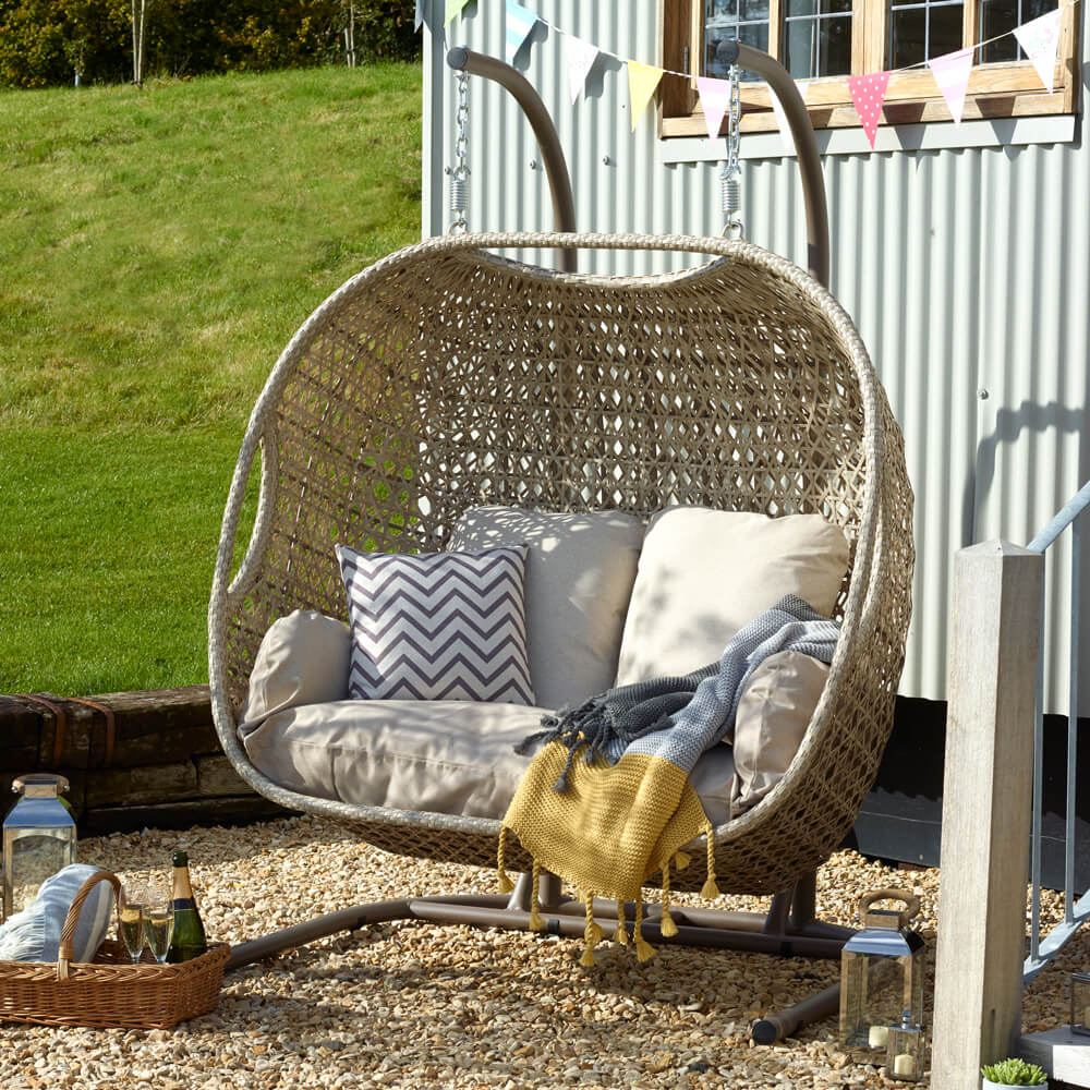 Bramblecrest Monterey Hanging Garden Chair With Cushions On Gravel with a basket containing a bottle of champaign and 2 glasses in front of it